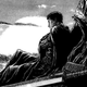 The swordsman Guts leans against a withered tree stump, his giant sword lying beside him.